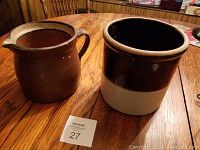 Photo showing both antique crocks side by side on a wooden table, one brown with handle and spout and the other two-tone brown and beige.