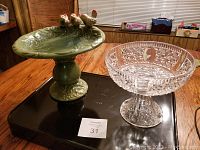 Photo of two compote dishes on a wooden table: one clear cut crystal with geometric patterns, one green ceramic with bird figurines on rim