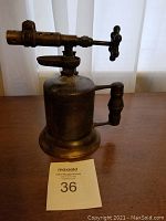 Side view of antique brass torch showing cylindrical body, wooden handle, and horizontal nozzle with control valve.