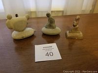 Three sculptures displayed on a wooden surface near a window curtain: two stone sculptures and one smaller brown resin sculpture.