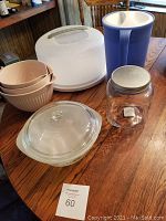 Kitchenware displayed on wooden table including pink mixing bowls, blue and white pitcher, clear glass casserole dish, glass storage jar, and white plastic cake carrier.