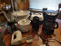 Photo of the lot showing all kitchen appliances on a wood table including the crock-pot, blender, hand mixer, coffee maker, electric skillet, food warmer, and can opener.