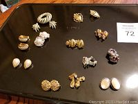 Photo showing a black tray with 14 pairs of vintage clip-on earrings in varied styles and colors, showcasing gold, pearl, enamel, and rhinestone details.