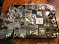 Group photo showing most earrings in clear ziplock bags laid out on black tray with some loose pairs