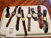 Nine men's wristwatches laid out flat on a white tray showing variety of bands and dial styles.