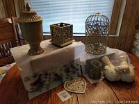 Photo of the lot showing ornate brass urn, decorative metal bird cage planter, ceramic lovebirds statuaries, glass bird tray, glazed pottery small urn, and heart-shaped welcome sign on a wooden table with a white speckled cloth underneath.