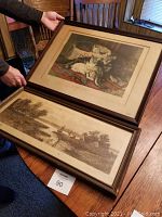 Photo showing both framed pictures with glass and wooden frames; one picture features cats on a patterned rug, the other a landscape with sailboats.