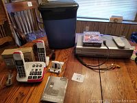 Full view of lot items on wooden table including Panasonic cordless phone system with base and handsets, black paper shredder, Sylvania VHS-DVD player with remote and John Wayne DVDs.