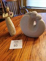 Both figurines side by side on a wooden table: a rounded stone sculpture mother and child and a taller cream-colored Willow Tree Angel figurine
