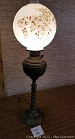 Full view of the antique Victorian banquet lamp showing the round cherry blossom hand-painted glass globe on a dark brass ornate base and twisted stem with electric cord visible.