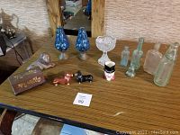 Complete view of all items on a wooden table showing bottles, candle holders, salt & pepper shakers, Toby mug, lion figurine, ink well, compote and tin matchbox holder