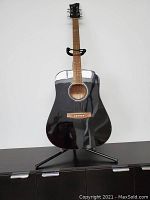 Front view of black acoustic guitar on stand showing body, strings, fretboard, bridge, and overall shape.