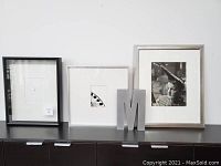 Four assorted wall art pieces including black and silver framed photos and a metallic letter M, displayed on a cabinet.