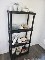 Photo of four-tier black shelving unit holding assorted clear glass plates, white ceramic cups, ramekins, small containers, and wooden utensils.