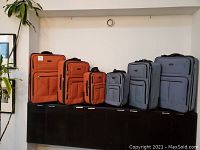 Photo of two complete luggage sets (three pieces each) side by side, orange set and blue-grey set, showing size differences and front pockets.
