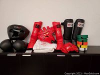 Full view of gloves, shin and arm guards, headgear, belts, and folded gi arranged on black cabinet.