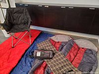 Four sleeping bags spread out (two rectangular red and blue, two barrel-shaped red/grey), black foldable chair, picnic blanket, cooler bag placed on top of sleeping bags.