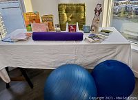Wide view of table showing large blue exercise ball, half blue balance ball, purple yoga mat, green cushion, various books, angel-themed decor items including bookends and figurines.