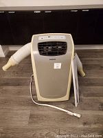 Front view of the portable air conditioner unit showing the vent grille, power cord, and attached ventilation hose on the left side, beige and gray coloring.