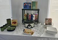 Full display of green stoneware dinnerware, glasses, cookbooks, digital Crockpot, water bottles, and white plates and bowls arranged on table and shelving.
