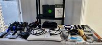 All gaming items arranged on a white table including Xbox, Playstation 2, Wii Fit balance board, multiple controllers, wires, and accessories.
