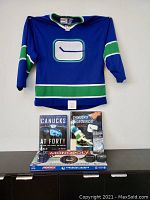 Full view of the large CCM Vancouver Canucks retro hockey jersey with prominent logo, plus the two Canucks books, Monopoly game, and collectible pucks lined up beneath.
