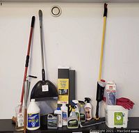 Full view of cleaning supplies and equipment arranged on black cabinet including broom, dustpan, mops, cleaning solutions, tiki lamp and pink gloves