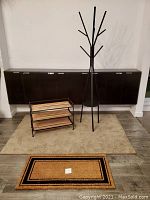 Photo showing all items together: tan/dark beige area rug beneath black shoe rack and black modern coat/hat rack, smaller entry rug in front.