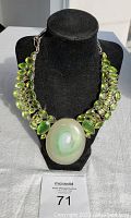 Full view of necklace on black stand showing large polished green oval center stone surrounded by smaller green faceted stones in silver-tone setting.