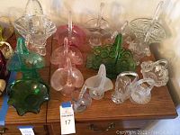 Top-down view of a collection of 12 vintage glass baskets in various colors and styles arranged on a wooden surface, showing details of handles, rims and decorative patterns.