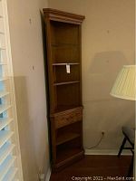 Full view of tall corner shelving unit positioned in a corner by a side lamp and wall outlet. Shows the shape and finish of the piece.