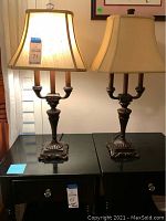Pair of bronze color candelabra lamps with cream fabric shades on black side tables under warm lighting showing slight marks on shades.