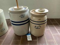Photo showing two ceramic butter churns side by side on tiled floor, one with a wooden churn stick and crown emblem, the other with blue stripes and markings.