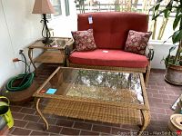Full set showing wicker patio settee with red cushions, wicker glass-top coffee table, wicker glass-top side table with lamp on top, and two decorative pillows.