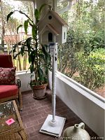 Full view of the wooden decorative church birdhouse on a tall stand with square base, placed on a back porch beside a plant and furniture.