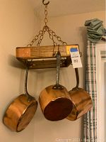 Overall view of 3 copper pots hanging on wooden square pot rack suspended by metal chain against beige wall.