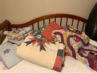 Photo of multiple folded and layered vintage quilts on a wooden slat bed frame, showing varied colors and patterns including floral and geometric designs.