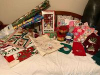 Overview of assorted Christmas table linens, gift bags, wrapping paper, and holiday items arranged on a bed and chair.