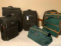 Front view showing three black Delsey suitcases plus green garment and duffle bags on a bed