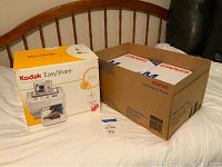 Kodak EasyShare G600 printer dock box and large Staples box of photo paper packages on a bed.