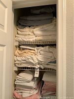 Closet shelving with multiple piles of folded assorted sheets and bedding items in various colors including gray, cream, beige, pink, and white.