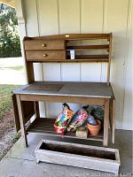 Full view of wooden potting table showing metal top inset, drawers, slatted lower shelf with gardening materials and planter box at front