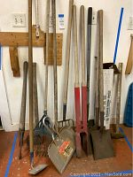 Photo showing the full set of garden tools standing against a wall. Visible tools include shovels, rakes, post hole digger, axe, and box of plastic sheeting. Tools have wooden handles and metal heads with some red paint on post hole digger.