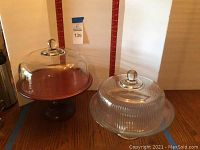 Two pedestal cake stands with clear glass domes shown side by side for size comparison and condition.