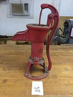 Side view of red cast iron water pump with manual pump handle and spout, sitting on a wooden table under an air conditioner.