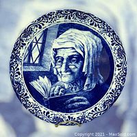 Front side of the Boch Freres Delft collector plate showing elderly woman reading book in blue and white motif with floral border.