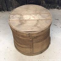 Top and side view of circular wooden cheese box with lid in place on concrete floor showing construction and wood coloration.