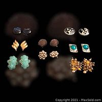 Top view of eight pairs of vintage clip-on earrings arranged in two rows on a black surface, showing diverse designs.