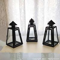Three black metal outdoor candle lanterns shown standing side by side on a table in front of lace curtains.