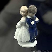Porcelain figurine of a girl and boy standing back to back on oval base, with girl dressed in white dress and boy in blue outfit, both with arms crossed or hands folded, detailed face and hair, gloss porcelain finish.
